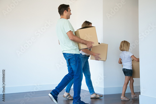Father holding cardboard box and going to corridor after wife and daughter. Cheerful young family with kid relocating in new house or apartment together. Mortgage, relocation and moving day concept