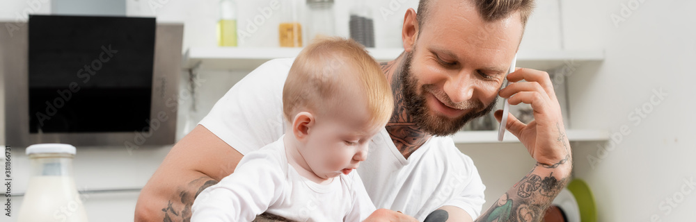 horizontal image of young man talking on smartphone in kitchen during breakfast with infant son horizontal image of young man talking on smartphone in kitchen during breakfast with infant son