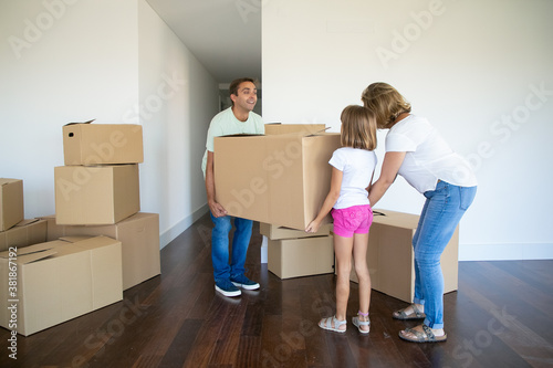 Happy parents and girl carrying box into new empty flat together. Full length. Real estate purchase concept © Mangostar