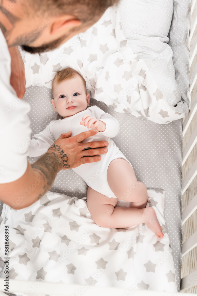 overhead view of father touching infant son lying in crib in baby romper overhead view of father touching infant son lying in crib in baby romper