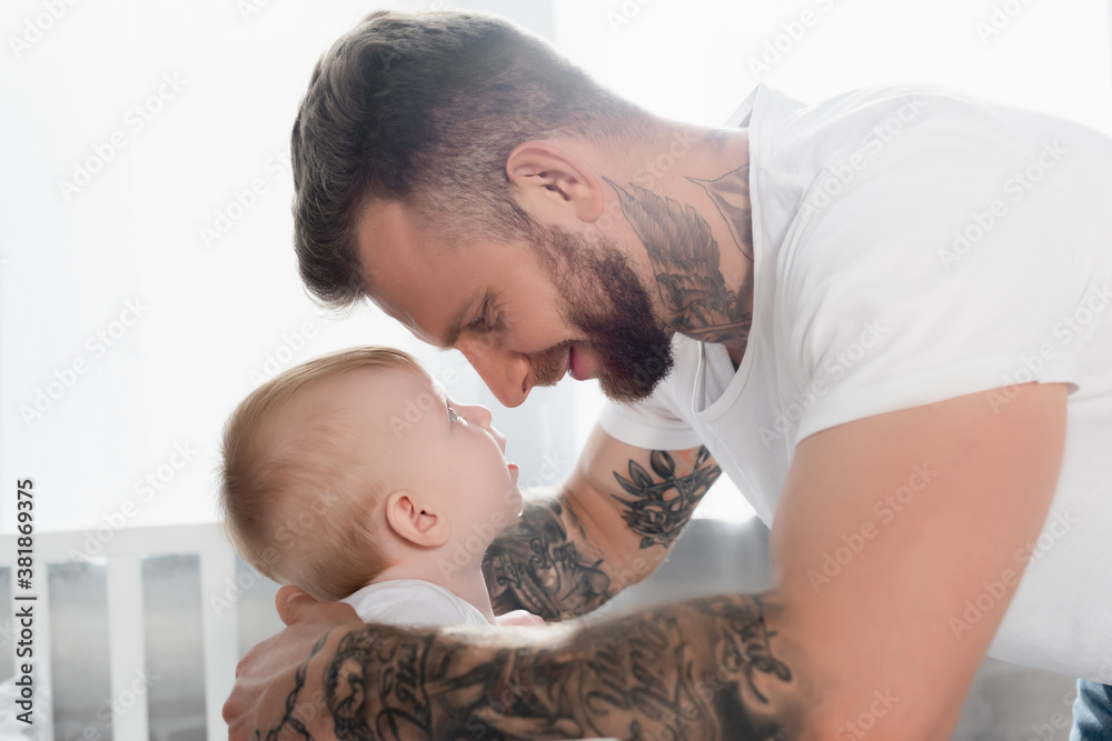 side view of joyful tattooed man and baby boy looking at each other face to face