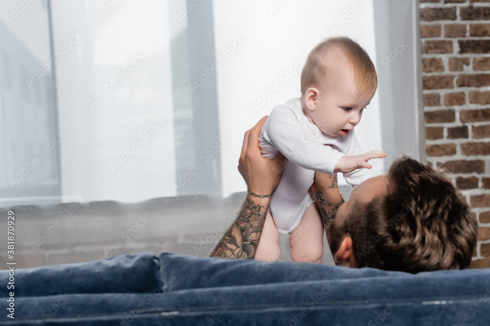 selective focus of tattooed man holding baby boy while sitting at home selective focus of tattooed man holding baby boy while sitting at home