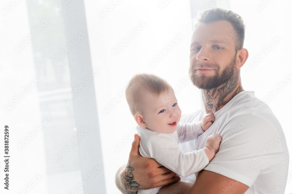 young tattooed man looking at camera while holding joyful infant son
