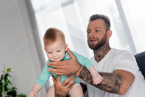 selective focus of bearded tattooed man holding baby boy in bedroom