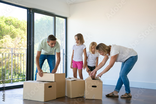 Happy family with two children unpacking boxes in new home. Caucasian parents with lovely blonde daughters standing in empty room with balcony. Mortgage, relocation and moving day concept