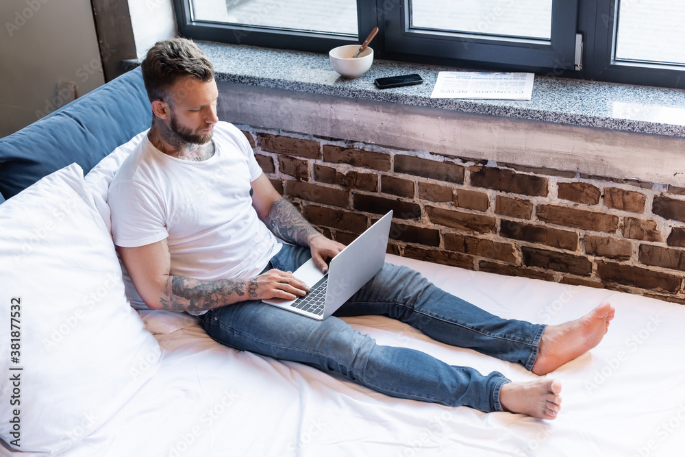 tattooed freelancer in jeans and white t-shirt using laptop on bed near newspaper, smartphone and bowl on windowsill
