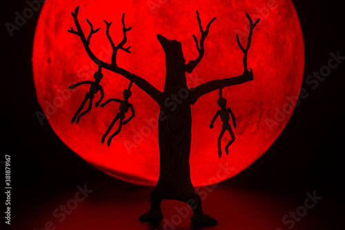 The silhouette of a tree standing dead with a man hanged dead to the bone. There's a red full moon in the background. Halloween horror concept.