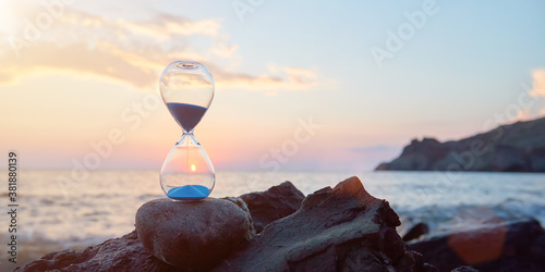 Hourglass with blue falling sand inside. Ocean landscape and golden hour. Time fly and need in rest concept, copy space