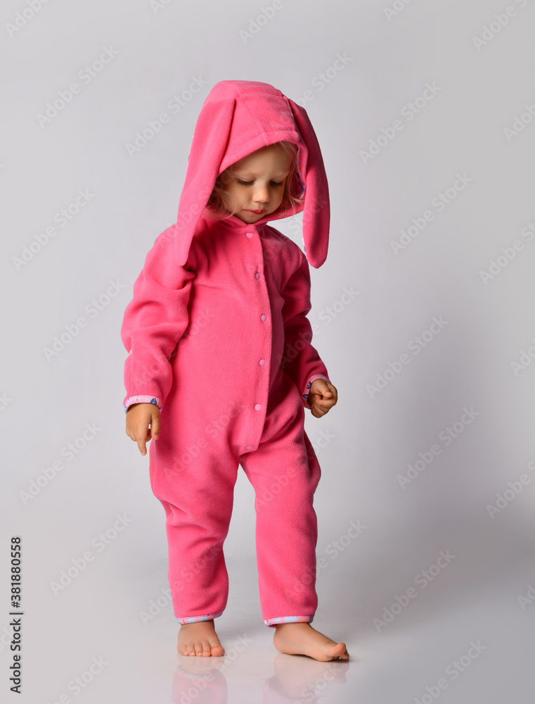 Pensive barefoot cute blonde baby girl in pink warm jumpsuit with hood and bunny ear walks slowly looking down Pensive barefoot cute blonde baby girl in pink warm jumpsuit with hood and bunny ear walks slowly looking down