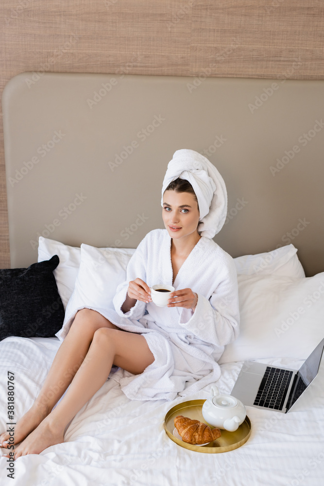 young woman in bathrobe holding cup of coffee near laptop and breakfast tray on bed young woman in bathrobe holding cup of coffee near laptop and breakfast tray on bed