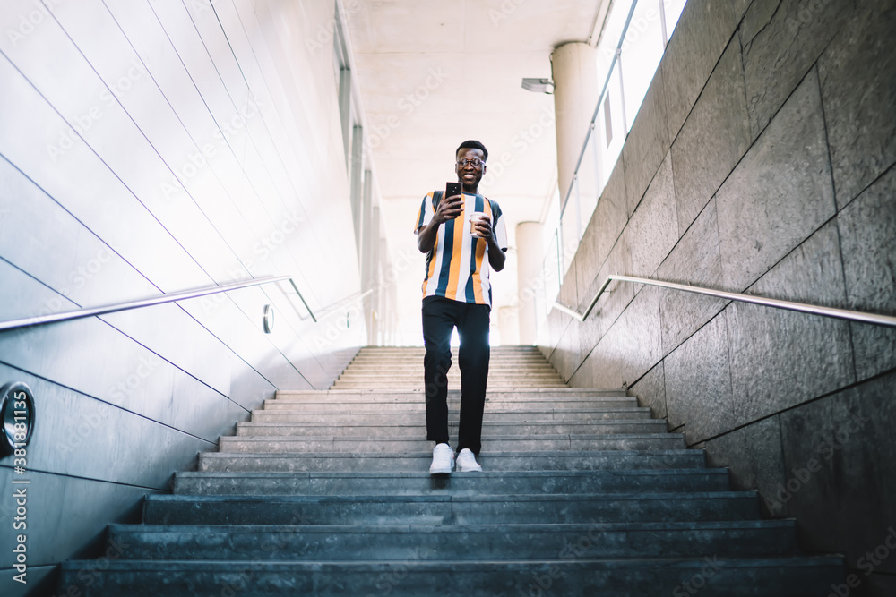 Positive black man walking downstairs and messaging on smartphone Positive black man walking downstairs and messaging on smartphone