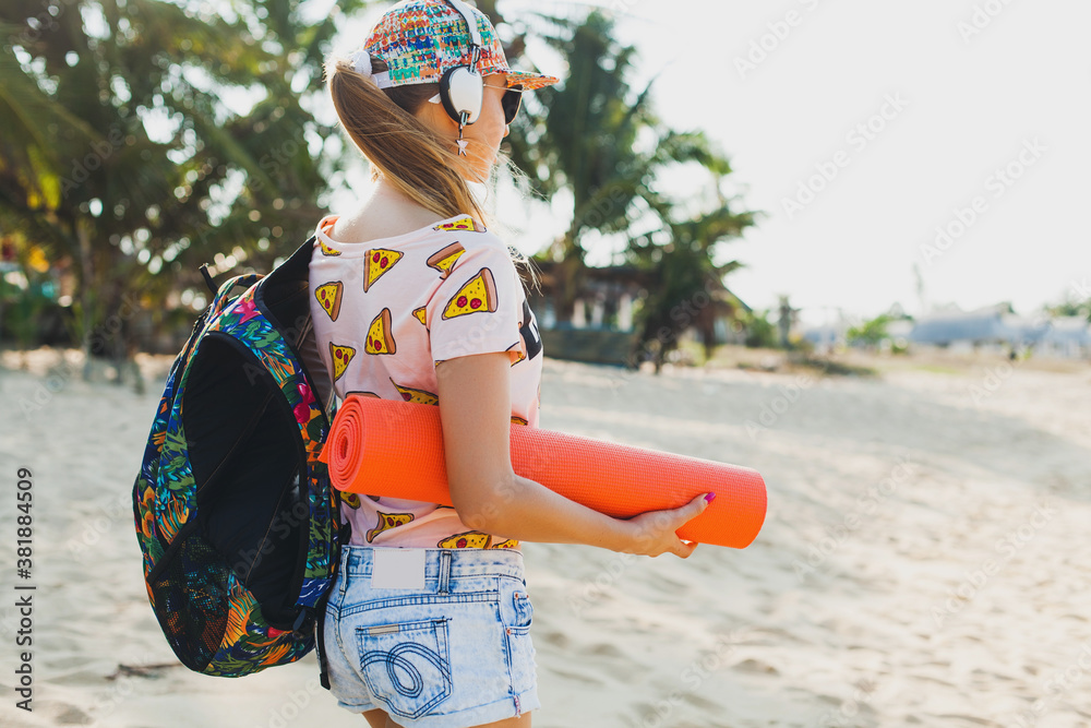 young beautiful woman walking on beach holding yoga mat, hipster sport swag style, denim shorts, t-shirt, backpack, sunny, summer weekend