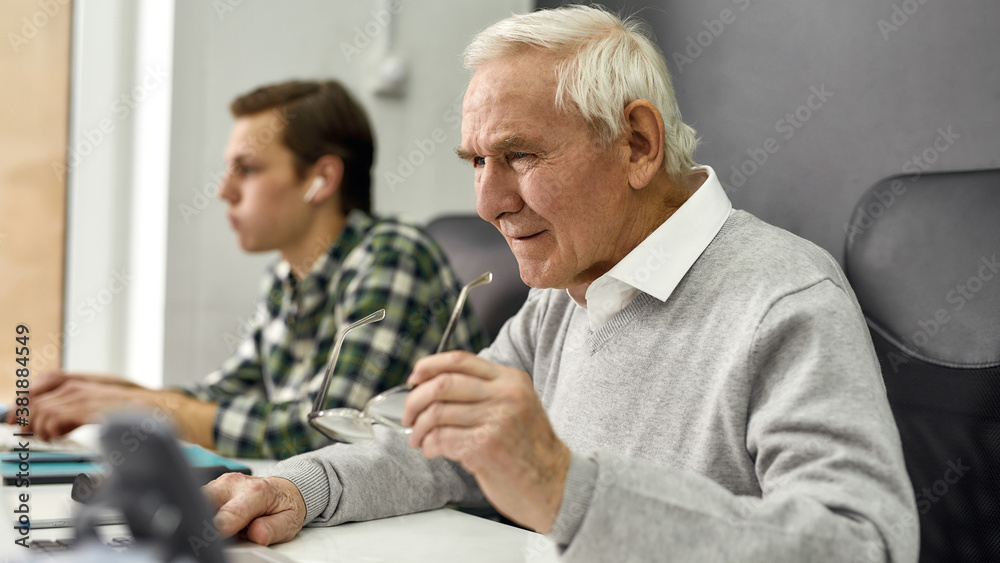 Close up of aged man, senior intern looking focused while using laptop, sitting at desk, working in modern office with young colleague