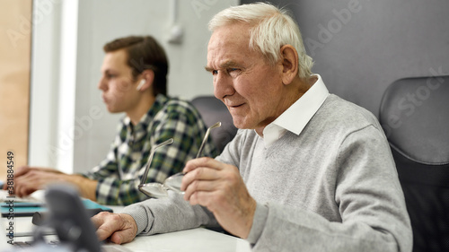 Close up of aged man, senior intern looking focused while using laptop, sitting at desk, working in modern office with young colleague