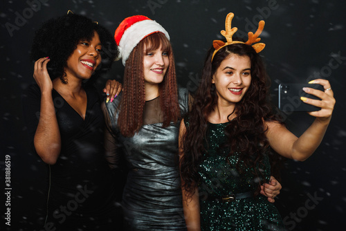 A group of female friends of different nationalities take a Christmas selfie in the studio. Young women in santa hats and deer antlers are having fun together