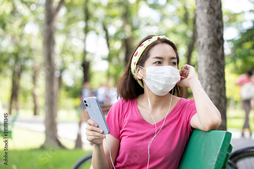 Woman wearing a face mask sitting using smartphone for listening music outdoors on a bench in a lush green park. concept of the coronavirus or Covid-19 pandemic.