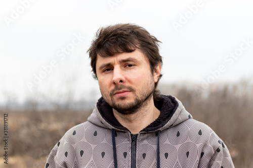 A handsome bearded man with disheveled hair against a blurred background. Man in a jacket with a hood