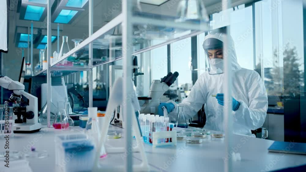 Beautiful Female Medical Scientist Wearing Coverall and Face Mask Using Micro Pipette while Working with Test Tubes. Vaccine, Drugs Research and Development Laboratory with Modern Equipment Beautiful Female Medical Scientist Wearing Coverall and Face Mask Using Micro Pipette while Working with Test Tubes. Vaccine, Drugs Research and Development Laboratory with Modern Equipment