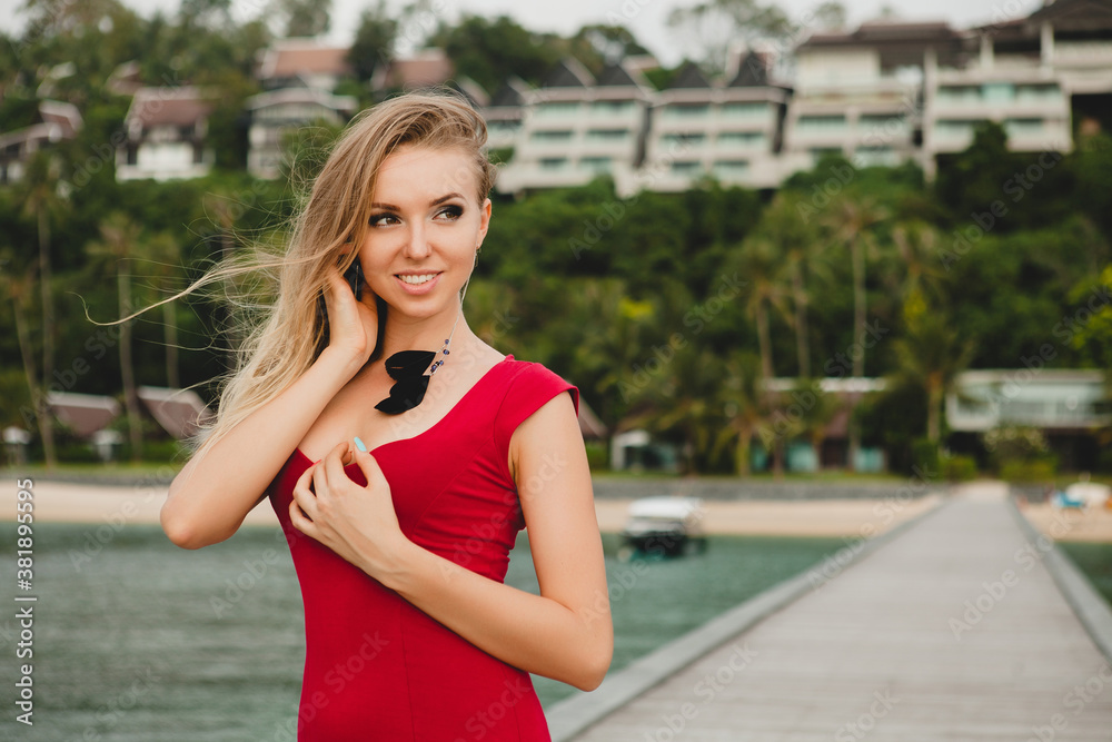 young beautiful attractive woman standing alone on pier in luxury resort hotel, summer vacation, red long dress, blond hair, sexy apparel, tropical beach, seductive, sensual, smiling