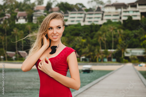 young beautiful attractive woman standing alone on pier in luxury resort hotel, summer vacation, red long dress, blond hair, sexy apparel, tropical beach, seductive, sensual, smiling