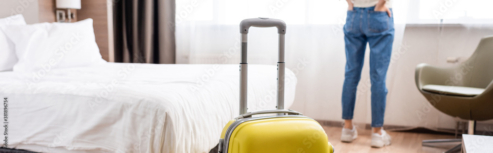 selective focus of yellow luggage near woman standing in hotel room, horizontal crop - obrazy, fototapety, plakaty 