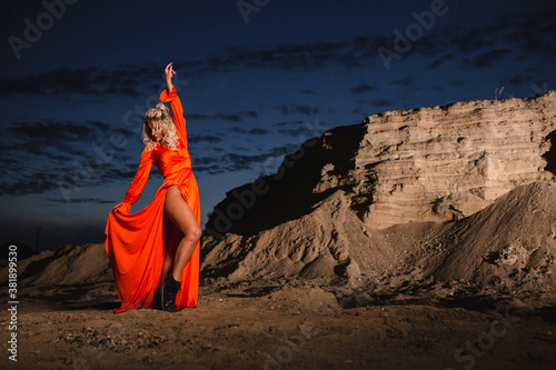 Woman in orange dress posing near the sand hill at the background of sunset
