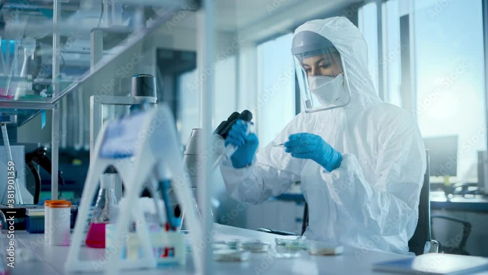 Beautiful Female Medical Scientist Wearing Coverall and Face Mask Using Micro Pipette while Working with Petri Dish. Vaccine, Drugs Research and Development Innovative Laboratory Modern Equipment 