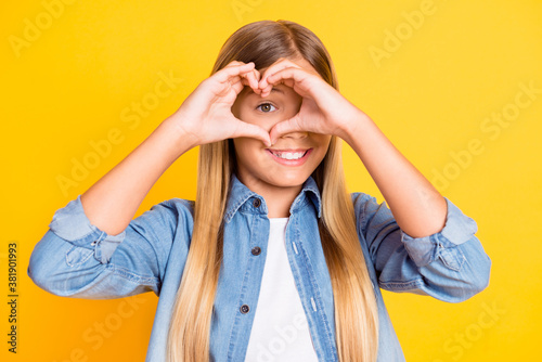 Photo portrait of pretty schoolgirl showing heart with her hands looking throw wearing denim shirt smiling isolated on bright yellow color background