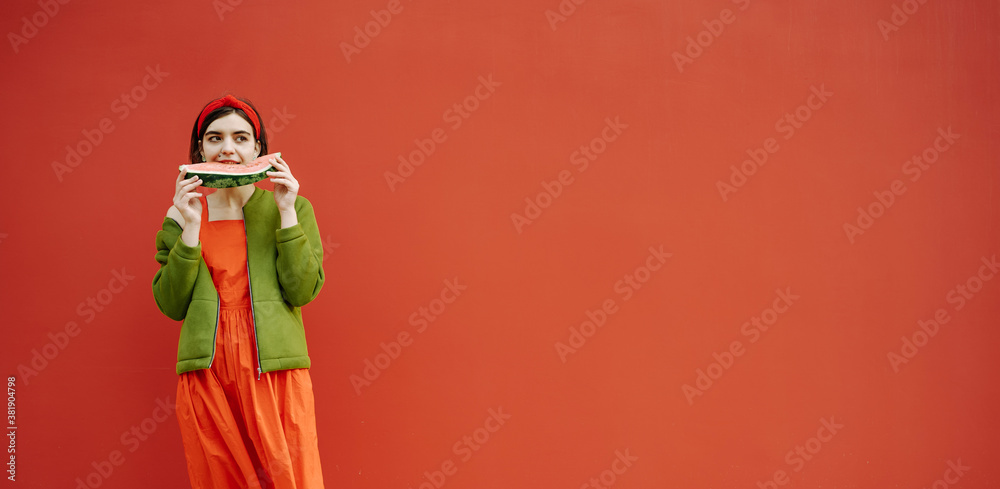 Long red banner. Young brunet girl eating watermelon Cool stylish red dress green jacket trendy clothes Funny mood fashion concept Bright emotional Horizontal composition Beautiful hair accessories 