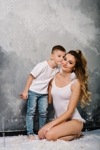 Smiling mom with son sitting on the floor. Fashion models plays with feathers. Happy family portrait in casual style clothes. Nice family wearing jeans isolated on grey wall. © Serhii