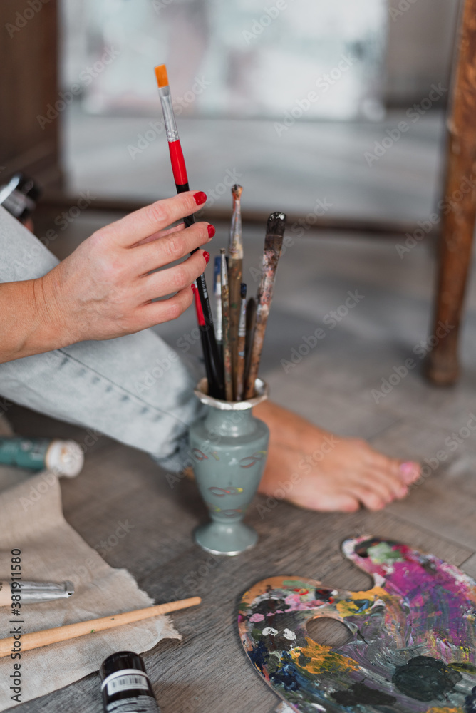 Obraz premium female hand holds a brush for painting. there is a vase with different tassels on the floor, next to it is a palette for stirring flowers
