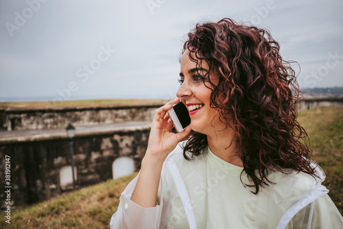 A beautiful young woman in a white rain jacket smiling and talking on the phone in a rainy day in the north of Spain.Technology and lifestyle concept.