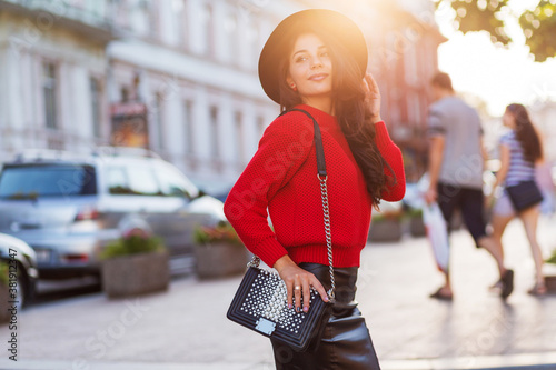 Outdoor fashion street style image of seductive brunette woman in autumn casual outfit walking in sunny city . Red knitted pullover, black trendy hat, leather skirt. photo