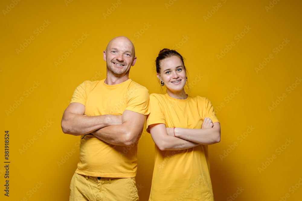 Obraz premium Funny, cute man and woman in plain bright clothes are standing with crossed arms and smiling benevolent, on a yellow background.
