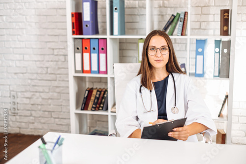 Focused young female doctor with a stethoscope on the shoulders sits at the desk and fills out an application form at office in clinic, looks at camera. Virtual appointment concept