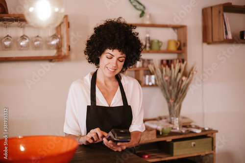 Smiling female cafe owner standing working with a card terminal. Woman with apron standing behind the bar counter of a modern restaurant. Concept small business owner, young entrepreneur.