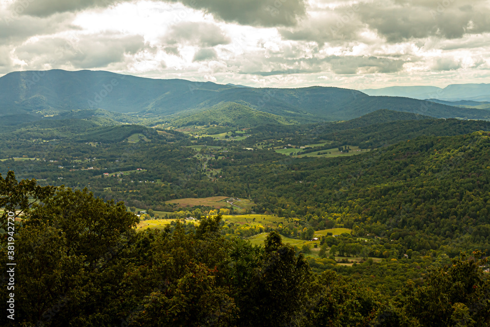 A scenic vista of Shenandoah Valley as seen from a scenic overlook by Skyline drive. Sunlight rays penetrate among clouds and there are small farms within a vast forest on valley floor.