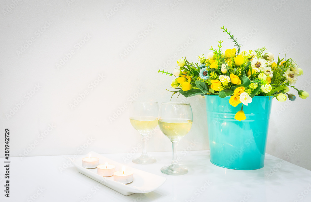 Romantic set up of burning candles, two white wine glasses and blooming flowers in bucket on white table and wall in background Romantic set up of burning candles, two white wine glasses and blooming flowers in bucket on white table and wall in background