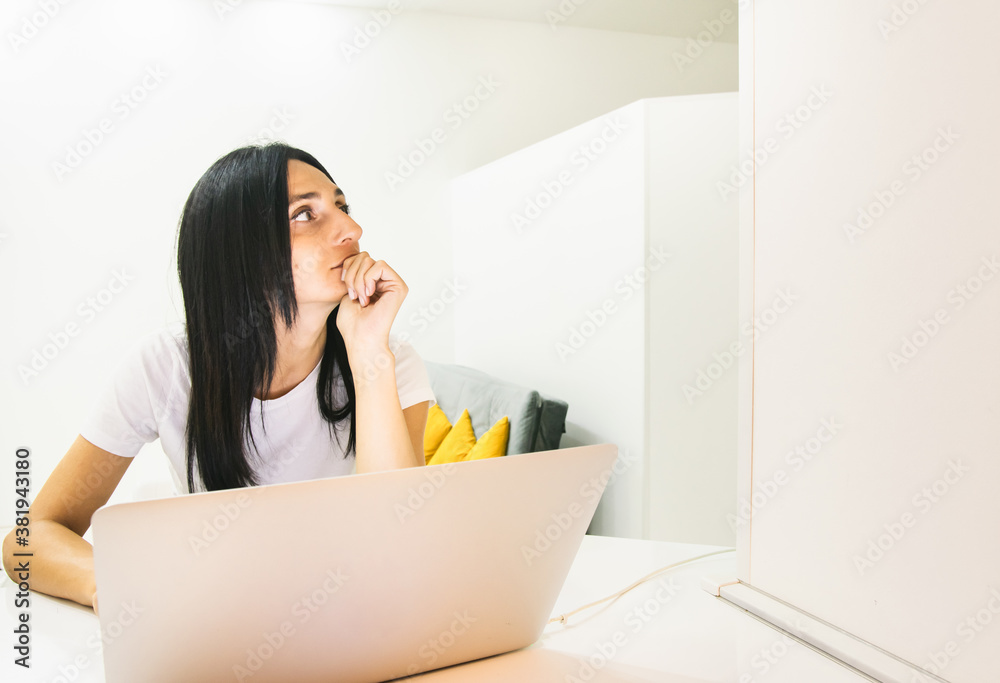 Young brunnete woman sits behind laptop and looks up being thoughtful. Concept of study and work from home. Young brunnete woman sits behind laptop and looks up being thoughtful. Concept of study and work from home.