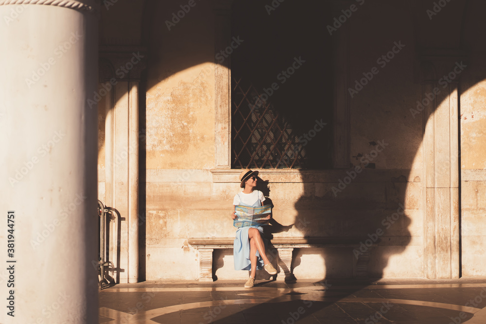 Cheerful woman holding map and sitting in sunny arched gallery