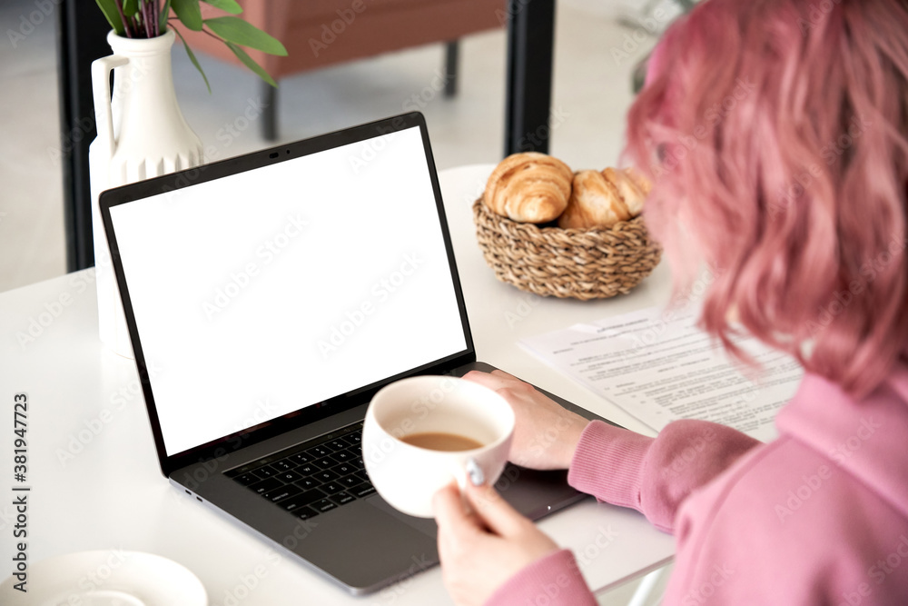 Hipster teen girl young woman student, worker sitting at kitchen table using laptop mock up white screen drink coffee learning online, working from home office, paying bills in app. Over shoulder view Hipster teen girl young woman student, worker sitting at kitchen table using laptop mock up white screen drink coffee learning online, working from home office, paying bills in app. Over shoulder view