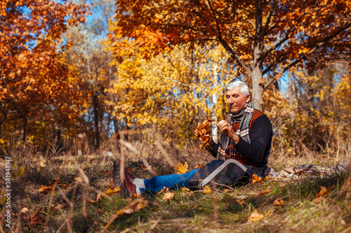 Senior alone man sitting in park picking yellow leaves. Retired man relaxing outdoors enjoying autumn nature