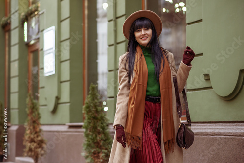 Φωτογραφία Happy smiling woman wearing trendy autumn, winter beige coat, hat, brown scarf, gloves, pleated skirt, with stylish faux leather bag, walking in street of European city