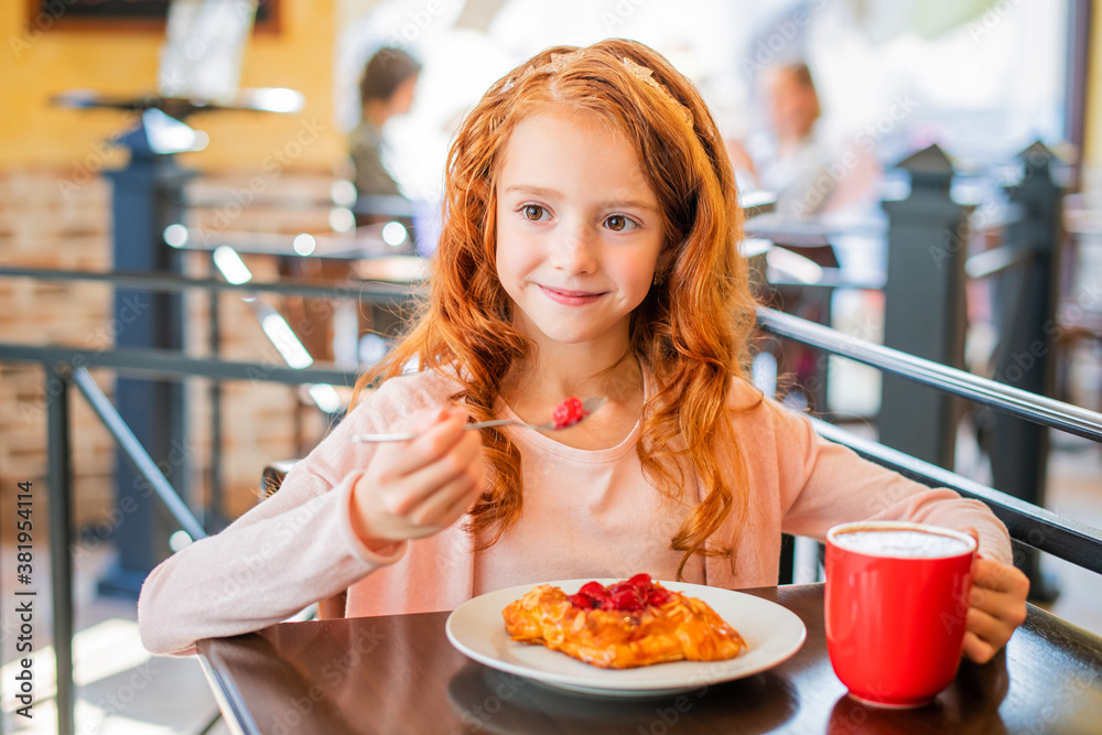 Little red-haired girl eating cake in cafe, smiling looking into the distance. Little red-haired girl eating cake in cafe, smiling looking into the distance.