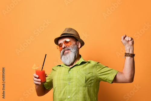 Gray-bearded man in hat, green shirt, sunglasses. Smiling, stretching himself, holding glass of juice, posing on orange background