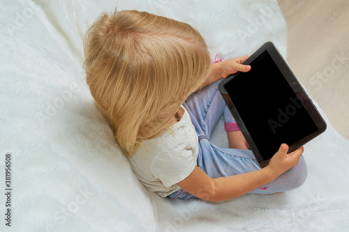 Blonde girl sitting on an armchair with a white fluffy blanket and holding a tablet in her hands, back view
