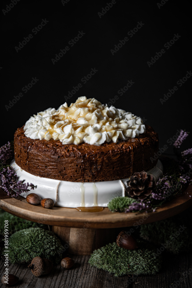 Homemade Carrot cake with ricotta cheese and honey frosting on wooden cake stand surrounded with moss, acorns, pines and with little white candle on foreground on black background.