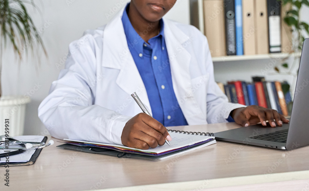 African american woman gp doctor wears white coat using laptop computer writes patient prescription notes records sits at desk. Black professional medic working at workplace in hospital. Close up view African american woman gp doctor wears white coat using laptop computer writes patient prescription notes records sits at desk. Black professional medic working at workplace in hospital. Close up view
