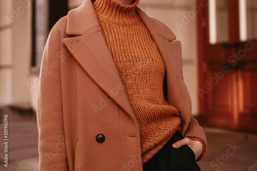 Young woman  with cropped head in the orange knitted cozy sweater and brown coat standing on the street. Outdoor portrait in daylight. Autumn casual fashion style  photo