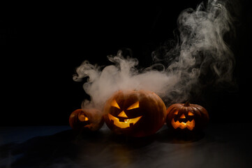 Three creepy halloween steaming pumpkins with a carved luminous smirk on a black background. A handmade jack-o-lantern head with a candle inside in the dark among the fog. Trick or treat. Postcard.